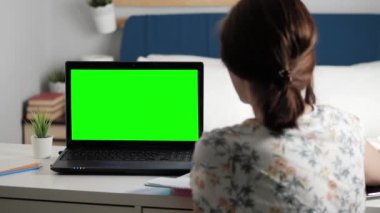 Kadın yatak odasında masa başında oturuyor, dizüstü bilgisayara yeşil ekrana bakıyor ve internetten biriyle konuşuyor, bazen not defterine not alıyor. Yakın plan.