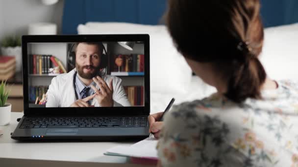 Dokter online verbinding. vrouw zit aan tafel in de voorkant van laptop en leidt gesprek met mannelijke arts in witte jas over internet video verbinding, meisje neemt notities in notebook. — Stockvideo