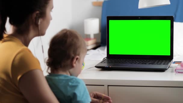 Mulher em fones de ouvido com criança senta-se na mesa no quarto, ela olha para a tela verde do laptop e fala com alguém sobre a conexão de vídeo da Internet, muitas vezes acena com a cabeça aprovadamente. Close-up — Vídeo de Stock