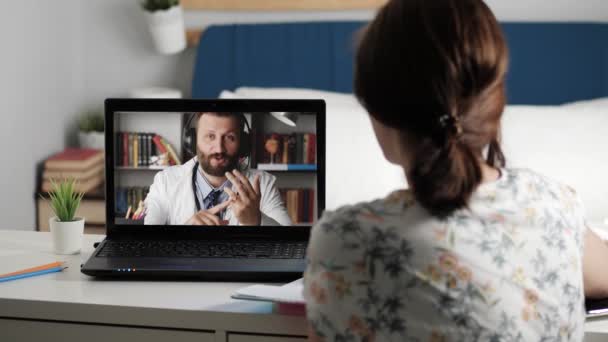 Lääkärin verkkoyhteys. Nainen istuu pöydässä kannettavan tietokoneen edessä ja johtaa keskustelua mieslääkärin kanssa valkoisessa takissa Internet-videoyhteyden kautta, tyttö tekee muistiinpanoja muistikirjaan. — kuvapankkivideo