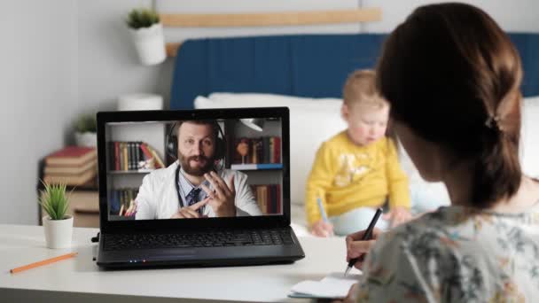 Enlace de vídeo de consulta en línea médico. Mujer mira a la pantalla del ordenador portátil y habla de la conexión de transmisión de Internet con el hombre de bata blanca. En el fondo está jugando un niño pequeño. Primer plano — Vídeos de Stock