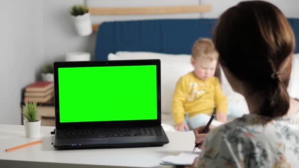 Mulher se senta na mesa no quarto, ela olha para a tela verde do laptop e fala com alguém através de comunicações de vídeo na internet, às vezes tomando notas no caderno. No fundo é criança pequena. Close-up — Vídeo de Stock