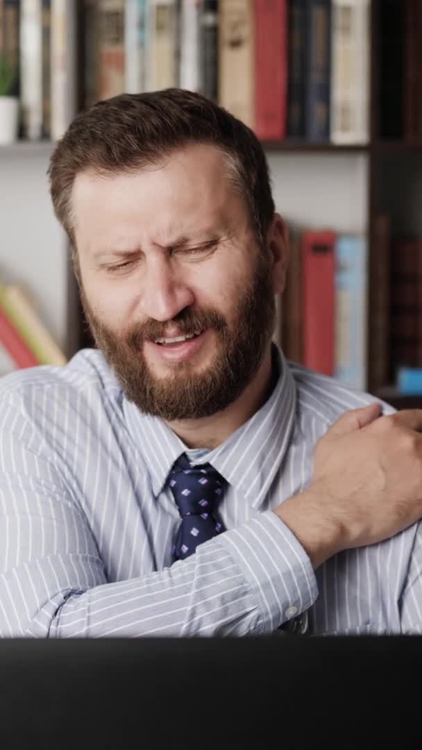 Shoulder pain. Vertical orientation man on workplace experiencing pain and discomfort in shoulder, he touches his shoulder with hand and stretches. Sprain, joint injury, sedentary lifestyle concept — Stock Video