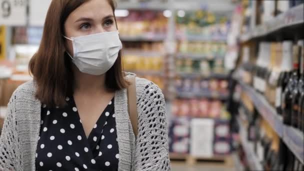 Kobieta w ochronnej masce chirurgicznej przechodzi przez supermarket z wózkiem obok półek z alkoholem i winem i patrzy na butelki. Zbliżenie — Wideo stockowe