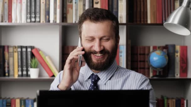 El hombre está hablando por teléfono y sonriendo. Hombre barbudo positivo en el lugar de trabajo hablando emocionalmente por teléfono y escribiendo en el portátil. Primer plano — Vídeos de Stock