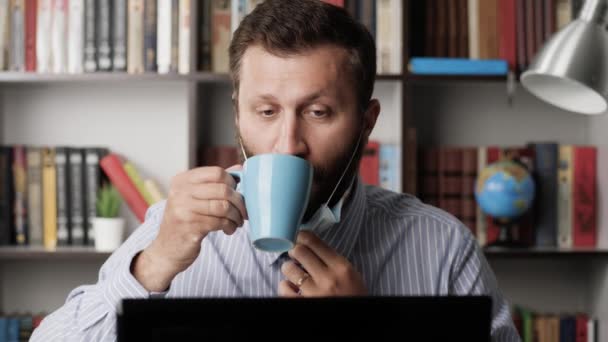 Mann mit OP-Maske am Arbeitsplatz tippt auf Laptop-Tastatur, arbeitet und trinkt Kaffee oder Tee aus blauem Becher. Pandemische Coronavirus covid-19-Konzept. Nahaufnahme — Stockvideo