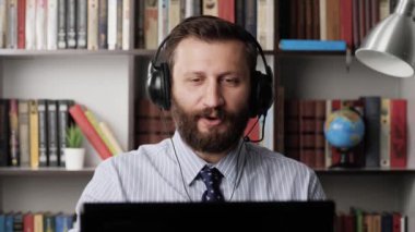 Adam video bağlantısından bahsediyor ve küfür ediyor. İş yerinde kulaklıklı sakallı bir adam olarak çalışmak ve laptopta duygusal olarak olumsuz konuşmak. Yakın plan.