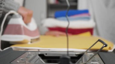 Kıyafetleri ütüleyen kız. Kadın eli ütüler sarı elbiseyi demirle ütüler sonra ütü masasına cihaz yerleştirir, ütü biter.