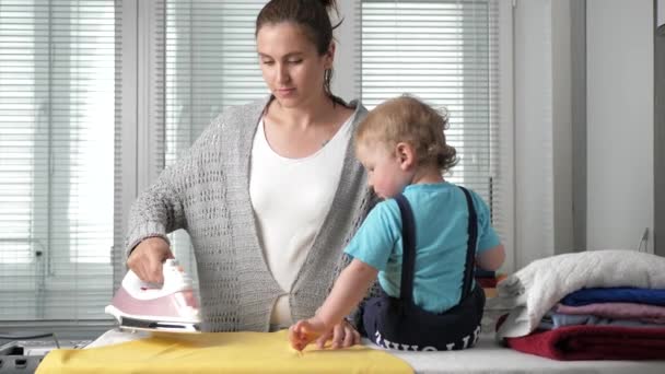 Holka žehlí oblečení. Mladá žena žehlí žluté šaty, malý chlapec sedí vedle žehlícího prkna — Stock video