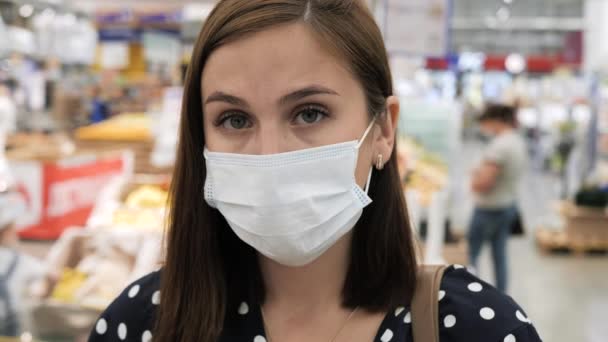 Žena v chirurgické ochranné masce je v supermarketu, dívá se na kameru, rozmazané maskované lidi chodí v pozadí. Coronavirus covid-19 koncept. Detailní záběr — Stock video