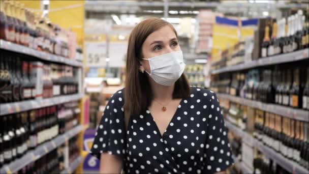 Kvinna i kirurgisk mask går genom snabbköpet förbi hyllor med alkohol, tittar på varor. Coronavirus covid-19 inköp av produkter koncept. Medelhög skott — Stockvideo