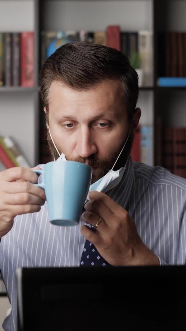 Verticale view man met chirurgisch masker op de werkplek typen op laptop toetsenbord, werken en drinken van koffie of thee uit blauwe mok. Pandemisch coronavirus covid-19 concept. Close-up — Stockvideo