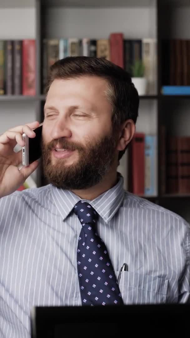 Чоловік розмовляє по телефону і посміхається. Вертикальний перегляд позитивний бородатий чоловік на робочому місці емоційно розмовляє по телефону і друкує на ноутбуці. Крупним планом — стокове відео