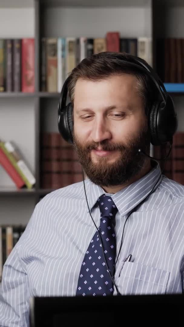 El hombre está hablando en la conexión de vídeo y sonriendo. Vertical ver hombre barbudo positivo en los auriculares en el lugar de trabajo emocionalmente hablando en video linc, se ríe y sonríe. Primer plano — Vídeo de stock