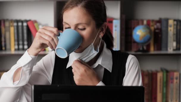 Жінка-працівник в хірургічній масці на робочому місці друкує на клавіатурі ноутбука, працює і п'є каву або чай з блакитного горнятка. Концепція пандемічного коронавірусу ковідо-19 . Крупним планом — стокове відео