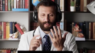 Doktor görüntülü arama. Beyaz önlüklü ve kulaklıklı bir erkek doktor iş yerindeki iş yerinde hasta ya da meslektaşlarıyla uzaktan iletişim kuruyor, bir şey anlatıyor ya da talimat veriyor.