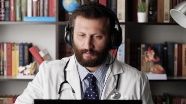 Doktor görüntülü arama. Hastane ofisindeki iş yerindeki kulaklıklı erkek doktor hasta ya da meslektaşlarıyla uzaktan iletişim kuruyor, dinliyor ve yazılı notlar alıyor.