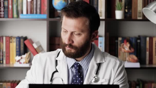 Doktor pracuje v počítači a pije kávu nebo čaj. Mužský lékař v bílém plášti na svém pracovišti v nemocniční kanceláři pracuje u počítače, píše na klávesnici a pije kávu nebo čaj z modrého hrnku — Stock video
