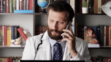 Doktor telefonda konuşuyor ve bilgisayardan not alıyor. Ofisindeki beyaz önlüklü erkek doktor cep telefonuyla konuşuyor ve aynı zamanda bilgisayar klavyesine bir şeyler yazıyor.