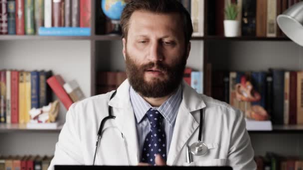 Tired doctor kneads his fingers and neck. Male doctor in white coat at his workplace in hospital office is typing on computer keyboard, is interrupted and begins to knead tired hands and neck — Stock Video