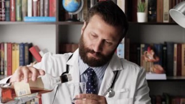 Üzgün doktor alkol içiyor. İş yerinde morali bozuk bir erkek doktor. Kahverengi alkolik. Konyak ya da viskiyi bardağa doldurup içiyor.