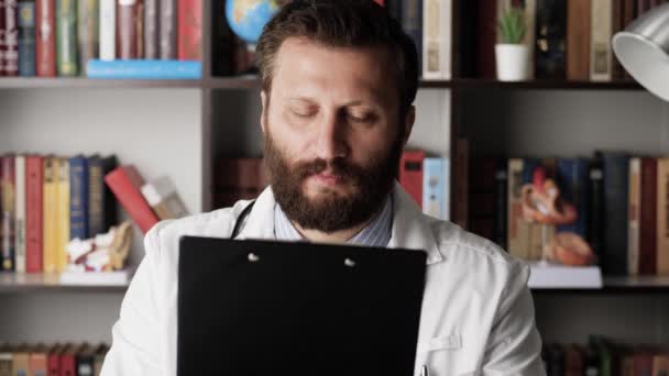 Доктор читає документи. Концентрований лікар чоловічої статі в білому пальто на робочому місці в лікарняному кабінеті тримає в руках документи або медичний запис і уважно читає текст — стокове відео