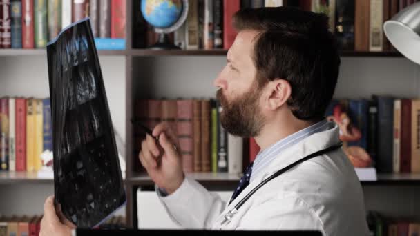 O médico lê a ressonância magnética. Médico do sexo masculino em seu local de trabalho no escritório do hospital detém ressonância magnética em suas mãos, examina-lo por muito tempo e faz anotações escritas para si mesmo — Vídeo de Stock