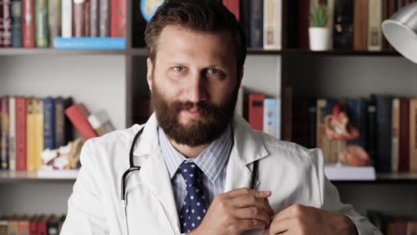 Doctorul se uită în cameră. Portretul unui doctor pozitiv de sex masculin în haină albă la locul de muncă în biroul spitalului care se uită la cameră și zâmbește — Videoclip de stoc