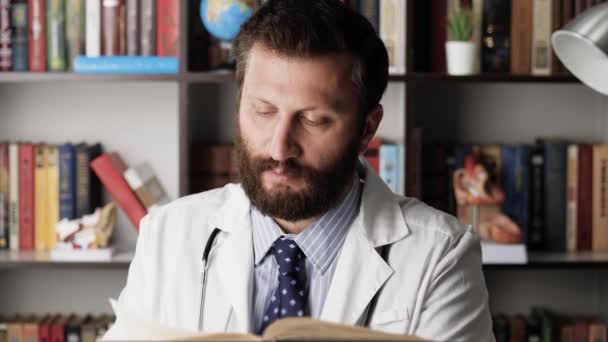 De dokter leest het boek. Man arts in witte jas op de werkplek in het ziekenhuis kantoor zit aan tafel en leest grote papieren boek, draait pagina — Stockvideo