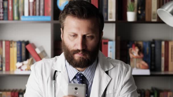 De dokter gebruikt telefoon. Man arts in witte jas op het werk in het ziekenhuis kantoor zit aan tafel en leest nieuws feed op de telefoon of schrijft bericht — Stockvideo