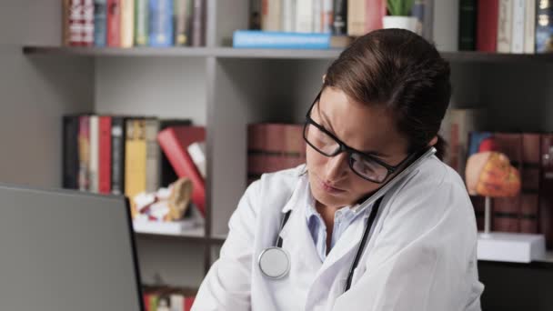 De dokter is aan het bellen en aan het typen op de computer. Vrouwelijke arts in witte jas op de werkplek houdt telefoon met haar schouder en met twee handen types op de computer toetsenbord — Stockvideo