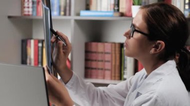 Doktor röntgen ya da MRI taraması okuyor. Ofisteki beyaz önlüklü kadın doktor röntgen veya MRI taramasına bakar ve notlar alır.