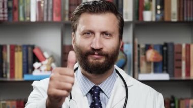Doktor parmak yukarı. Beyaz önlüklü arkadaş canlısı erkek doktor ve ofiste steteskop kameraya bakar ve elini kaldırır, başparmağını kaldırır, onaylayıcı bir şekilde başını sallar ve gülümser.
