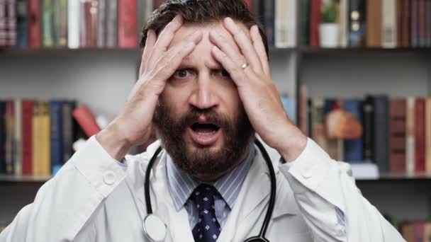 Horror doctor. Frightened excited male doctor in white coat and stethoscope in office looks at camera and covers his face with his hands — Stock Video