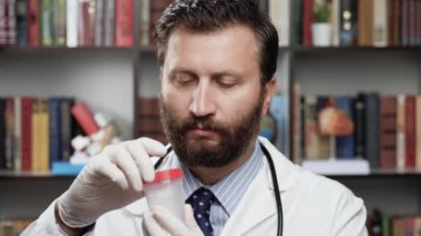 Test kavanozu. Beyaz önlüklü ciddi bir erkek doktor ve ofiste steteskop kameraya bakar ve analiz kabını kaldırır ve vidaları söker ve kameraya test yapmak için kavanozu gösterir.
