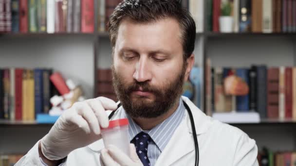 Pot d'essai. Médecin masculin sérieux en manteau blanc et stéthoscope au bureau regarde la caméra et ramasse le récipient d'analyse et dévisse le couvercle et montre le pot pour prendre des tests à la caméra — Video