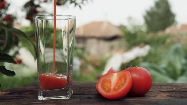 O suco de tomate derrama-se no vidro. O suco de tomate é derramado em vidro transparente contra o pano de fundo da casa de campo e da natureza, tomates vermelhos cortados frescos estão nas proximidades. Close-up — Vídeo de Stock