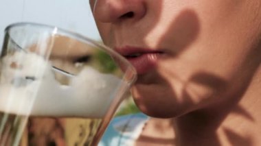 Kadın bira içiyor. Kadın elinin yan görüntüsü ağızlarına hafif bira ile cam getiriyor ve doğanın güneşli bir gününde içiyor. Yakın plan.