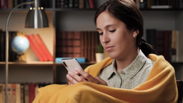 Woman uses phone in evening. Good mood girl is alone in evening or night in room wrapped in blanket uses smartphone, surfs Internet or makes online purchases — Stock Video