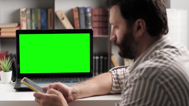 Mann sitzt am Tisch und schaut in Laptop grünen Bildschirm tippt er auf der Bildschirmtastatur des Smartphones und spricht über Internet-Online-Videoverbindung. Chroma Schlüssel Computerbildschirm Mann spricht auf Videoanruf — Stockvideo