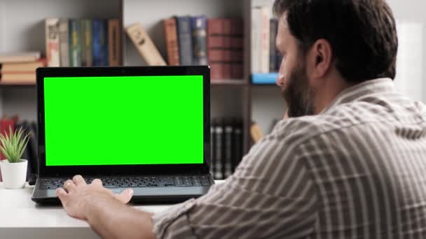 Mannen som sitter vid bordet i lägenhet eller kontorsrum tittar på bärbar grön skärm, han kör fingret på pekplattan och ibland trycker på knappen. Chroma nyckel datorskärm man arbetar på datorn — Stockvideo
