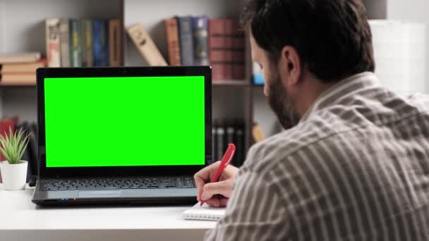 Hombre sentado en la mesa y mira en la pantalla verde del ordenador portátil, toma notas en el cuaderno mientras habla en línea conexión de vídeo por Internet. Chroma clave ordenador pantalla hombre hablando en videollamada — Vídeos de Stock