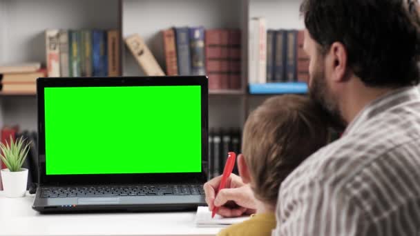 Homem e bebê sentado à mesa no quarto e olhando para a tela verde do laptop, o pai está falando na internet conexão de vídeo on-line e faz anotações. Chroma chave wom tela do computador falando em chamada de vídeo — Vídeo de Stock