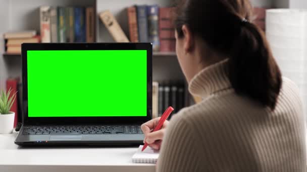 Žena sedí u stolu a dívá se na zelenou obrazovku notebooku, dělá si poznámky do notebooku a mluví na internetu on-line video připojení. Chroma klíč počítač obrazovka žena mluví na videohovor — Stock video