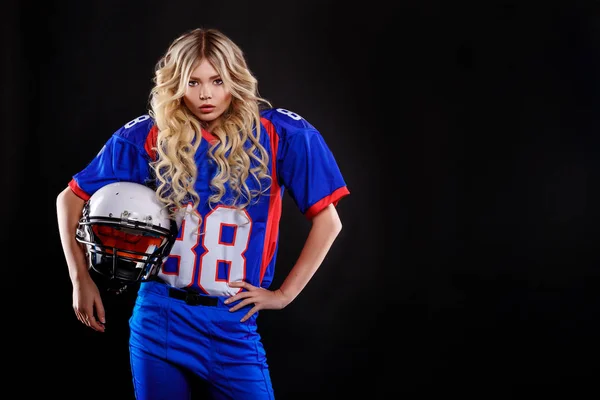athletic blonde posing as american football player on black background. Beautiful young woman wearing American football top holding ball.