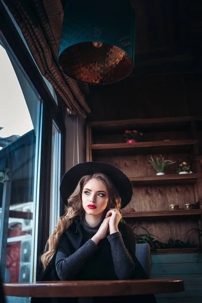 Mooie modieuze jongedame zitten in de buurt van grote venster in café en kopje koffie houden. Aantrekkelijke vrouw met stijlvolle zwarte hoed. — Stockfoto