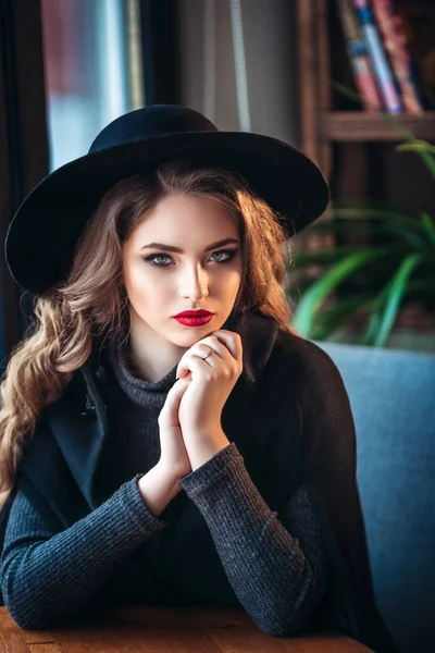 Junge schöne modische Dame sitzt in der Nähe eines großen Fensters im Café und hält eine Tasse Kaffee. attraktive Frau mit stylischem schwarzen Hut. — Stockfoto