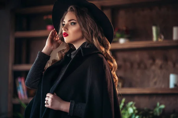 Jeune belle dame à la mode assise près d'une grande fenêtre dans un café et tenant une tasse de café. Belle femme portant un chapeau noir élégant . — Photo