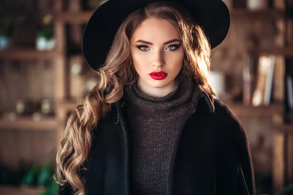 Junge schöne modische Dame sitzt in der Nähe eines großen Fensters im Café und hält eine Tasse Kaffee. attraktive Frau mit stylischem schwarzen Hut. — Stockfoto