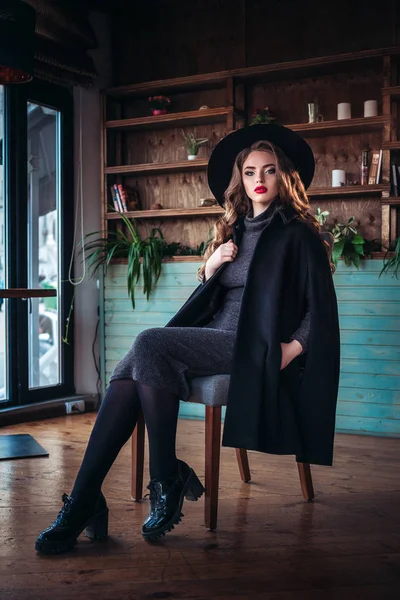 Mooie modieuze jongedame zitten in de buurt van grote venster in café en kopje koffie houden. Aantrekkelijke vrouw met stijlvolle zwarte hoed. — Stockfoto